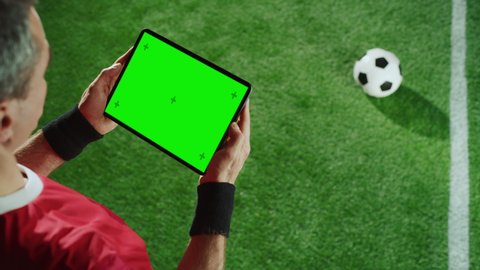 Professional Soccer Player Holding Digital Tablet Computer with Green Screen Chroma Key with Motion Markers. Elevated Top Down Over Shoulder Shot of Football Star Using Internet, Standing on Field
