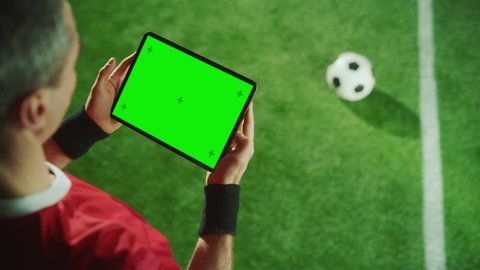 Professional Soccer Player Holding Digital Tablet Computer with Green Screen Chroma Key with Motion Markers. Elevated Top Down Above Shot of Football Star Browsing Content While Standing on Game Field