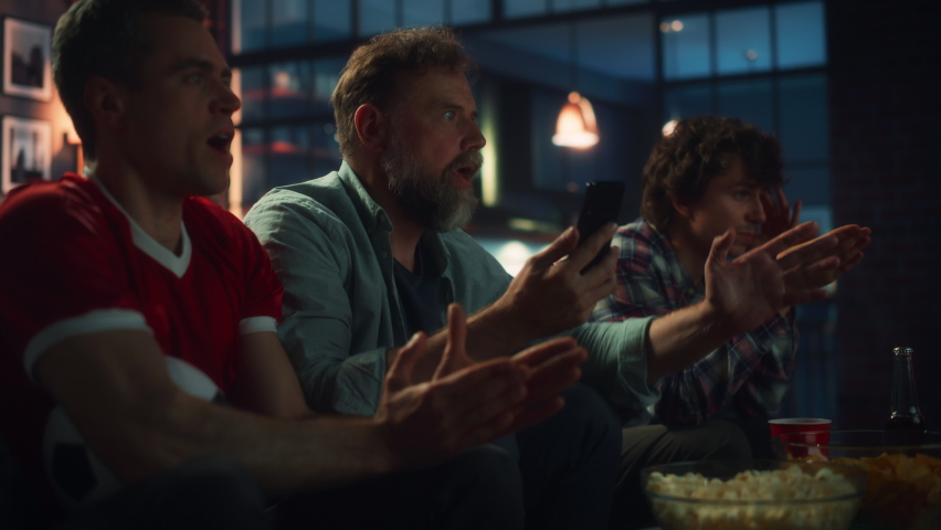 Night at Home: Three Soccer Fans Sitting on a Couch Watch Game on TV, Use Smartphone App to Online Bet, Celebrate Victory when Sports Team Wins. Friends Cheer Eat Snacks, Watch Football Play. | Shutterstock HD Video #1073965661