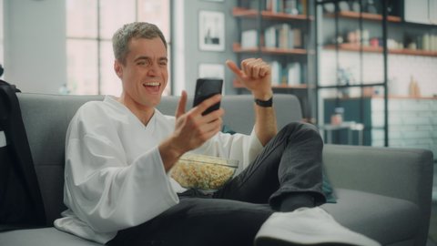 Handsome Man Sitting on a Couch Watches Ice Hockey Game on TV, uses Smartphone App for Score, Bet, Statistics, Celebrates Victory when Team Wins Championship. Happy Fan Watches Sport. Medium Shot