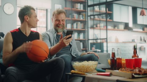 At Home Two Fans on Couch Watch Basketball Game on TV, Use Smartphone App for Checking Score, Online Bet, Celebrate when Team Wins Championship. Friends Watch Favourite Club Play. Wide Medium