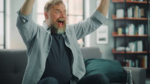 Charismatic Bearded Man Sitting on a Couch Watches Game on TV, Celebrates Sports when Team Wins Championship. Focused, Joyfully Intense and Authnetic Fan Cheers when Favourite Club Play