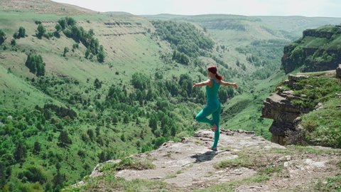 Athletic Girl in a Turquoise Jumpsuit Practices Yoga on the Edge of a Mountain. Slim Woman Workout Outdoors, Doing Balance Exercise against the backdrop of a Stunning Landscape. 