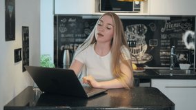 Positive woman with long blond hair smiling and talking with colleague while sitting at table and making video call on laptop during work in kitchen at home