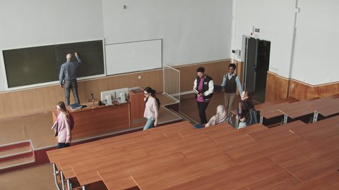High-angle slowmo of small group of young multiethnic university or high school students walking into big lecture hall while bold mid-adult professor writing topic of lesson on blackboard