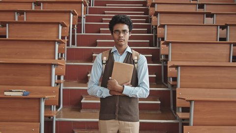 Zoom-in medium slowmo portrait of insecure Mixed-race university or high school student in eyeglasses with book in hands standing between aisle of long desks in lecture hall looking at camera
