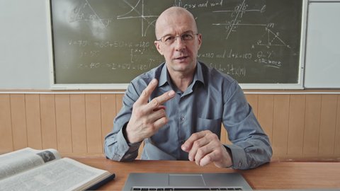 POV medium shot of confident male mid-adult professor teaching Maths speaking on camera while sitting by desk at blackboard in bright classroom