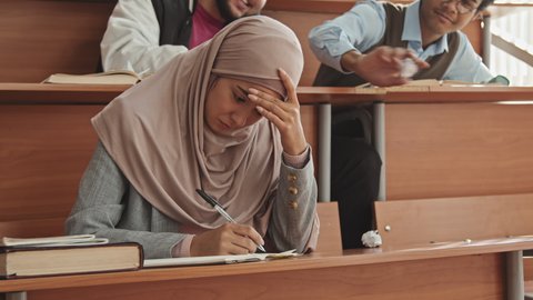 Medium slowmo of young female Muslim student in hijab sitting by desk crying in classroom while boys bullying her by throwing crumpled paper and laughing at her