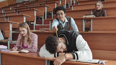 Medium shot of lazy male student in sports bomber jacket sleeping on desk in lecture hall or auditorium while his Mixed-race classmate poking at him with pencil to wake up as professor entering