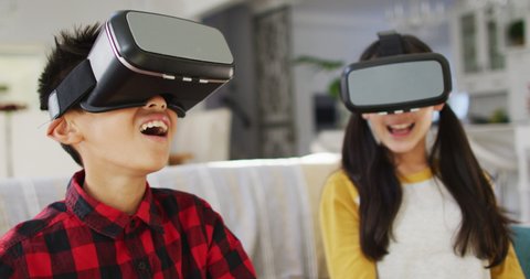 Happy asian brother and sister at home, sitting on couch in living room having fun using vr headsets. at home in isolation during quarantine lockdown.