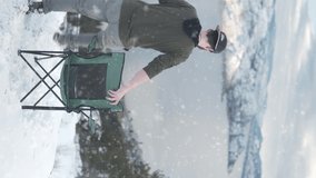 Vertical Video, Back of Young Man Sitting on Camping Chair on Snowfall With Lake View and Winter Landscape, Static Shot
