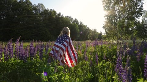 Unrecognizable blond woman wearing american flag walks in the meadow with lupin flowers at sunset. Memorial day concept. 