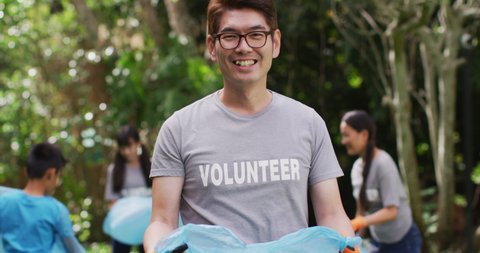 Smiling asian man wearing volunteer t shirt holding refuse sack for collecting plastic waste. eco conservation volunteers doing countryside clean-up.