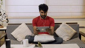 Indian man playing games on tablet PC with facial expressions. Young Asian guy excited with digital device while sitting at home indoor.lifestyle concept.Handsome male enjoy playing game in tablet.