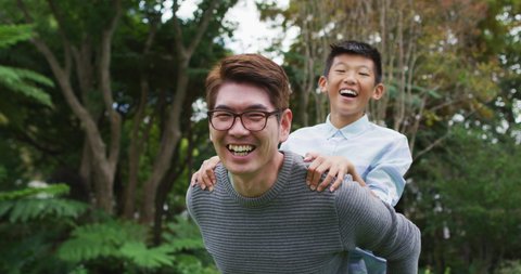 Smiling asian father piggy backing happy son having fun in garden together. happy family, at home in isolation during quarantine lockdown.