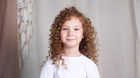 Preschooler little girl with long loose curly hair turns around and smiles slyly looking straight against grey background slow motion closeup