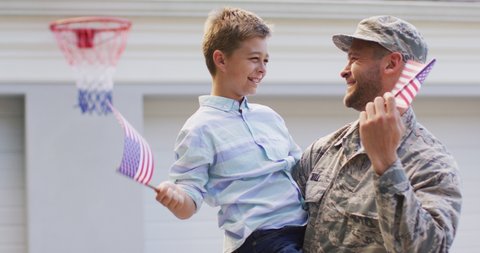 Happy caucasian male soldier carrying his smiling son, holding flags in garden outside their house. soldier returning home to family.