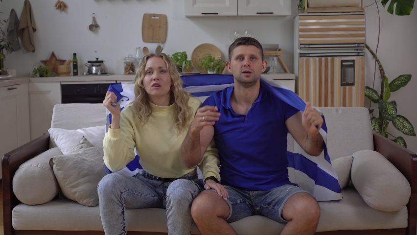Greek family couple of sports or football fans at home with the flag of Greece. Greece fans are watching TV.