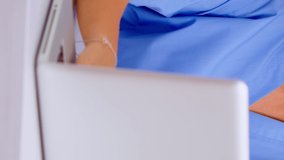 Vertical video: Close up of young medical assistant typing at laptop and raising head smiling at camera sitting in hospital office wearing blue uniform. Hospital healthcare worker, medicine clinical