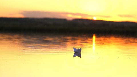 White paper boat floats in the water in the rays of the beautiful sun at sunset. Origami ship. Children's games, dreams of travel and discovery. Reliable business concept