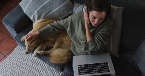 Smiling caucasian woman using laptop working from home with her pet dog on sofa next to her. pet companionship, domestic life and working from home concept.