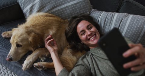 Smiling caucasian woman taking selfie with smartphone, stroking her pet dog on sofa next to her. pet companionship, domestic life and leisure time concept.