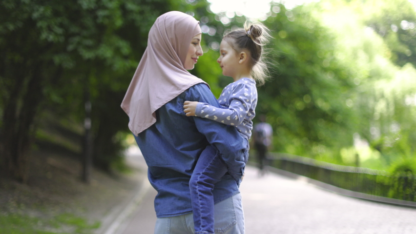 Young pretty happy Muslim mother in hijab, playing with her little adorable daughter in green park and holding her in arms. Slow motion of cheerful family, mom and kid girl, walking in the park Royalty-Free Stock Footage #1074026147