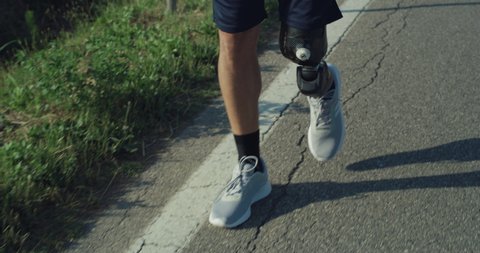 Cinematic close up of young man with disability uses artificial bionic high technology legs prosthesis walking on countryside road.Concept of persons with disabilities, active lifestyle, cyborg