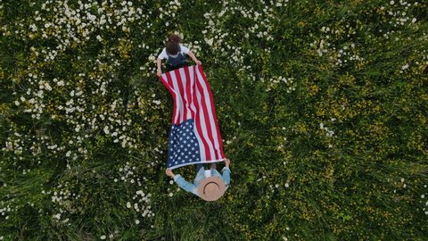 Drone shot of young playing mom with adorable girl kid holds proudly usa flag, demonstrates patriotism and freedom outdoors. Mother in hat with daughter standing on grass in middle of flowering field