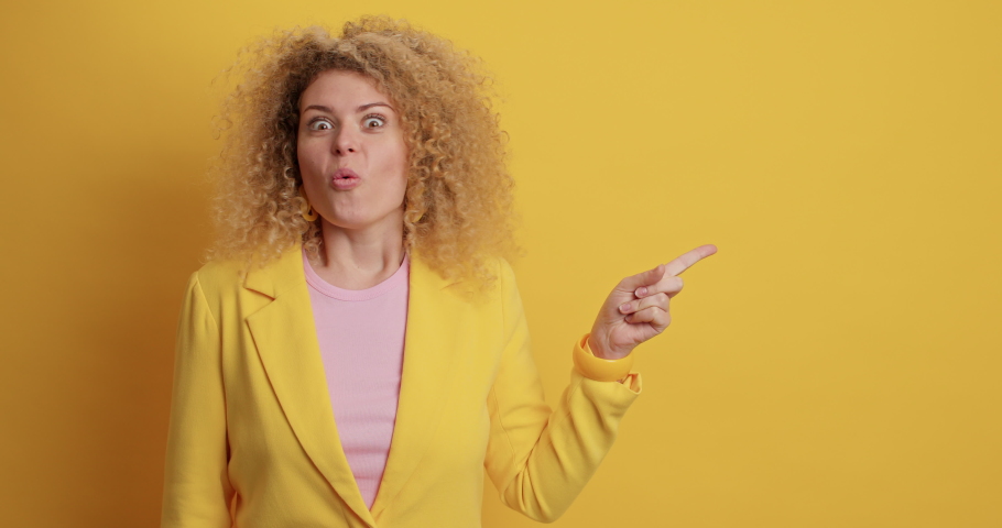 Amazed positive woman with curly hair has surprised glad expression says wow its great points away on blank copy space against yellow wall. Excited European female shows nice offer product on sale