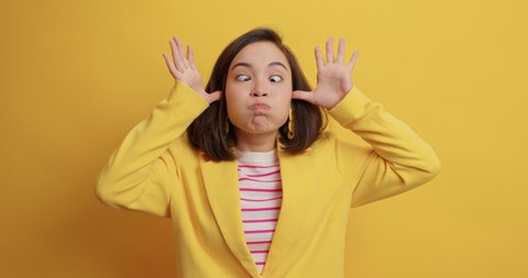Crazy funny brunette young Asian woman makes grimace foolishes around sticks out tongue dressed in formal clothing smiles joyfully isolated over yellow background. Human face expressions concept