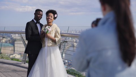 Happy African American newlyweds posing for photographer on bridge on sunny summer day. Smiling loving elegant bride and groom photographing outdoors on wedding day. Slow motion