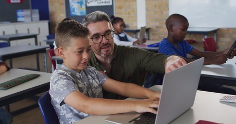 Diverse male teacher helping a schoolboy sitting in classroom using laptop. children in primary school.