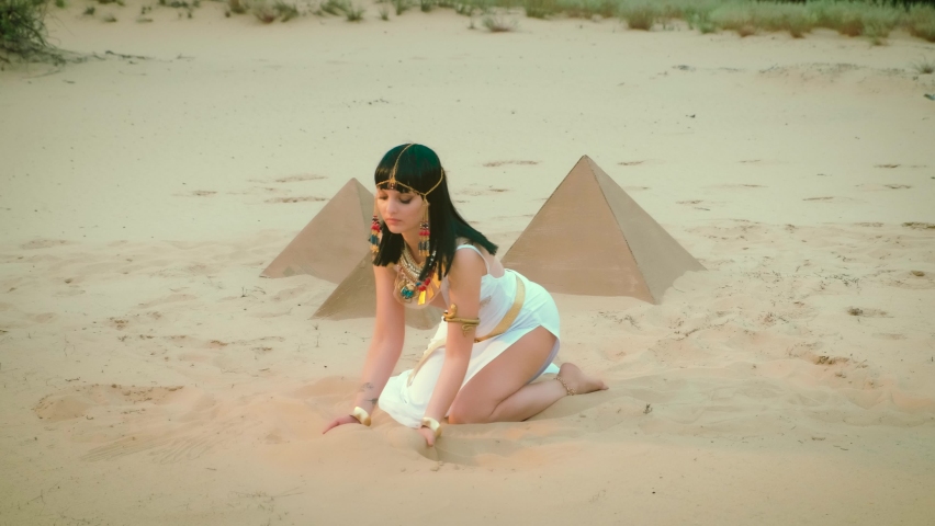 Beautiful sexy charming woman in image Egyptian pharaoh takes hands yellow sand, plays, conjures pours out. Hairstyle girl brunette princess decorated gold chain, necklace, bracelets. Pyramids, desert