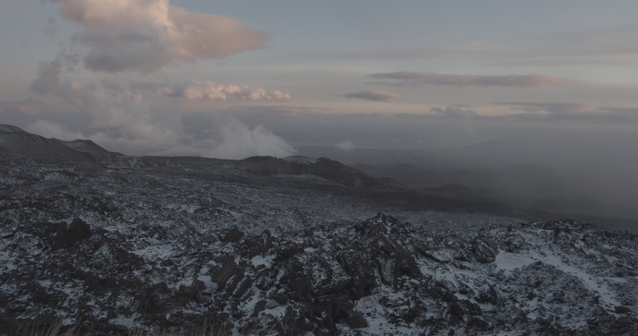 High and low hanging clouds over the Etna Vulcano in Italy which is partly covered with white snow over the black rocks on a windy dark colorful evening. Wide shot