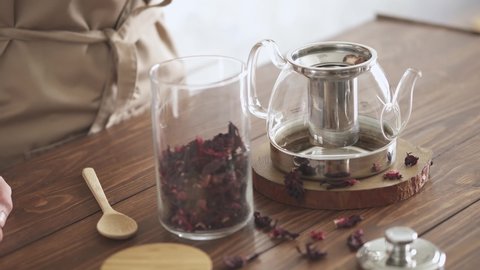 Red tea brewing process. Step one. A young housekeeper sprinkles dry hibiscus tea from a glass jar with a spoon. Pour hibiscus tea into a glass teapot. Making red herbal tea on a wooden kitchen table