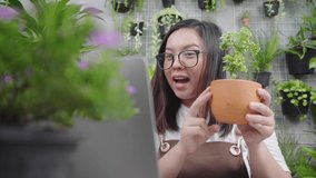 Happy Asian young woman selling plants online, holding plant product and using laptop to explain about the details to her customer by doing live on social media. Selling online concept
