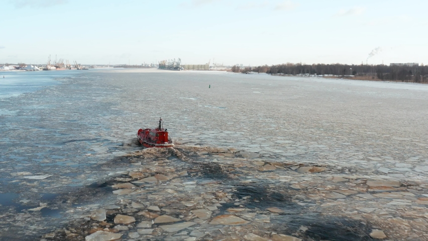 Aerial view follows small red ship sailing along drifting ice floes creating small ripple waves. Industrial port city in the background. Beautiful winter day. Forward movement. 4k footage.