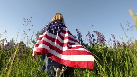 Unrecognizable blond woman wearing american flag walks in the meadow with lupin flowers at sunset. Low angle view. Memorial day concept. 