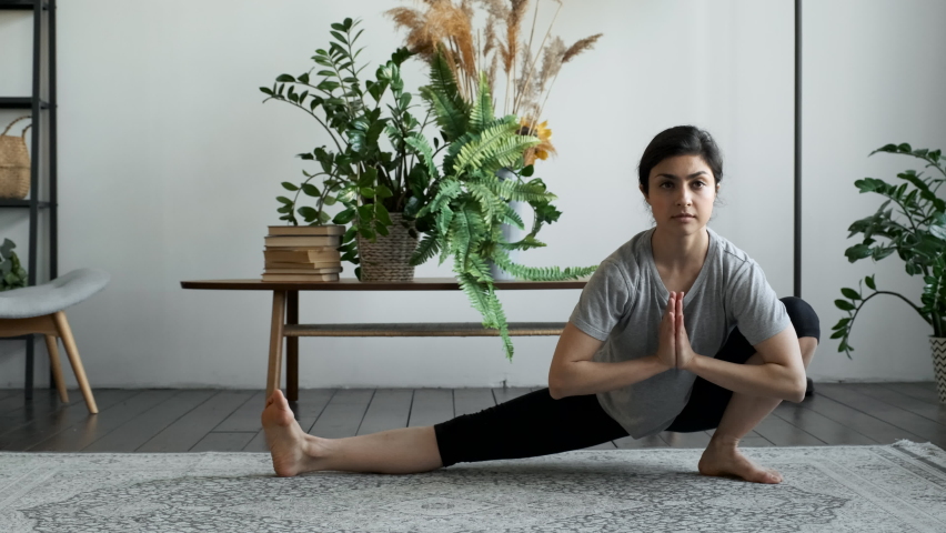 An Indian Woman Sits on the Floor and Prepares for a Yoga Workout, Doing Warm-Ups and Muscle Stretching, Squat Exercises to the Right and Left. Located at Home in a Cozy Room Royalty-Free Stock Footage #1074065363