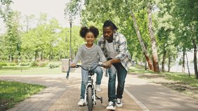 Father teaches his daughter to ride a two-wheeled bike. Childrens bike, a girl learns to ride for the first time. Happy Holidays African Ethnicity Family. - Powered by Shutterstock - Get 15% off with code: PIKWIZARD15