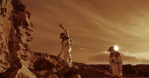 Low angle of people in spacesuits climbing on stones near cliff and looking away during colonization of Mars