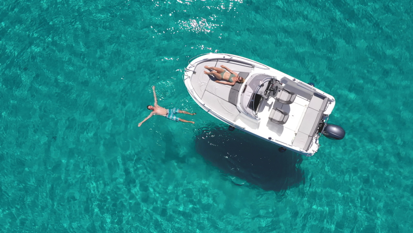 Aerial - Young couple enjoying their day on a boat on a beautiful turquoise sea. Man relaxes in the water by floating on the surface | Shutterstock HD Video #1074079130