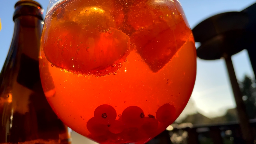 Orange alcoholic cocktail drink with wild berries against sunlight and blue sky in garden. Sun reflection in glass with ice cubes. Close up shot.