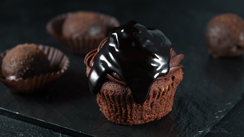 Pouring extra dark melted chocolate sauce over over a delicious, tasty cupcake in slow motion. Sweet biscuits, homemade pastry, on dark grey black slate background or texture