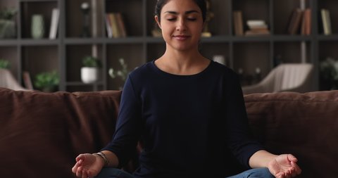 Indian ethnicity young pretty woman do meditation practice sit alone cross-legged on sofa in cozy living room with eyes closed. Stress free weekend, boost mindfulness training, visualization concept