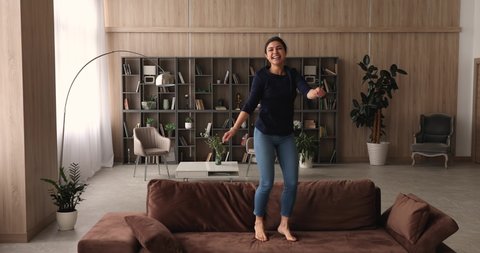 Cheery young Indian woman dance jumps on soft sofa in modern design cozy living room. Female carefree housewife listens music funny moving barefoot on couch. Enjoy weekend leisure, home hobby concept