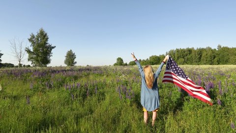 Blonde woman standing in meadow with her hands up holding American flag while other handshowing two fingers up in the peace or victory symbol. Independence day concept.