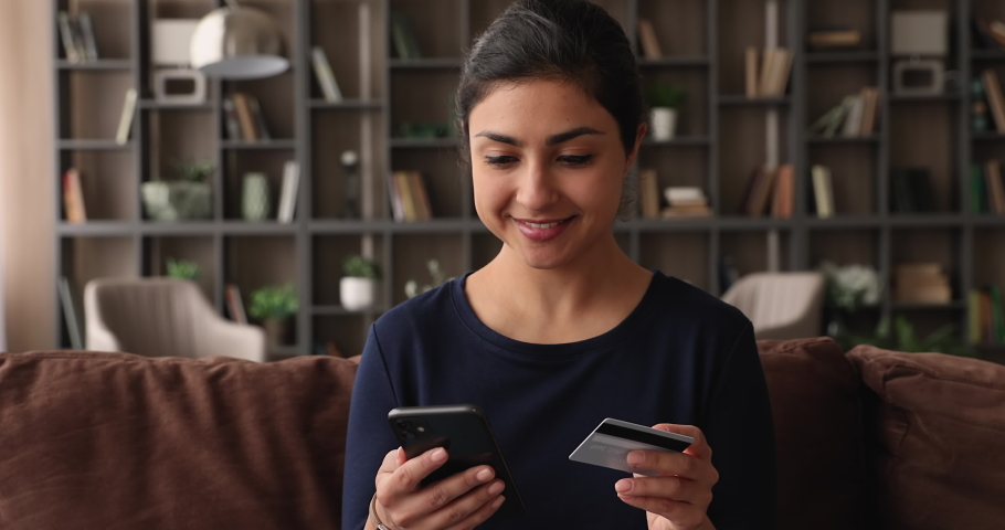 Happy young attractive Indian ethnicity woman sit on sofa holding smartphone and credit card, shopper making purchase online, use e-commerce retail web services buying fashion clothes goods for home | Shutterstock HD Video #1074110186