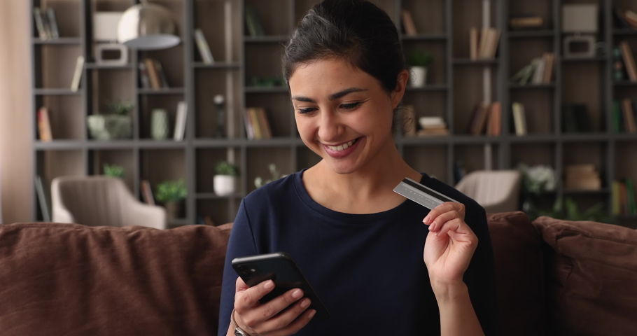 Happy young attractive Indian ethnicity woman sit on sofa holding smartphone and credit card, shopper making purchase online, use e-commerce retail web services buying fashion clothes goods for home Royalty-Free Stock Footage #1074110186